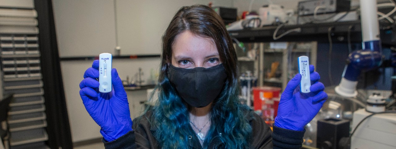 Amy Drexelius wearing a facemask and latex gloves holds up two testing devices in UC's Novel Device Lab.