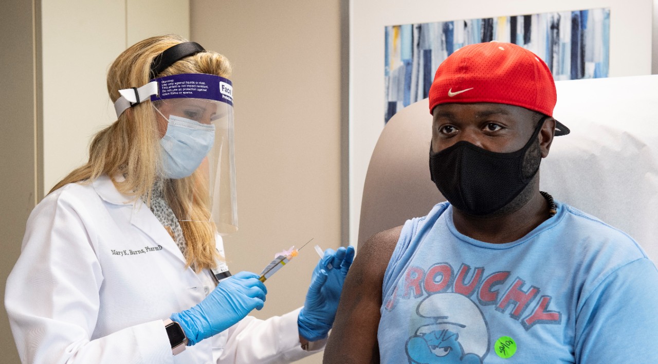 A pharmacist gives a vaccine shot to a Black man in a blue smurfs t-shirt wearing a black face mask and red baseball cap backwards