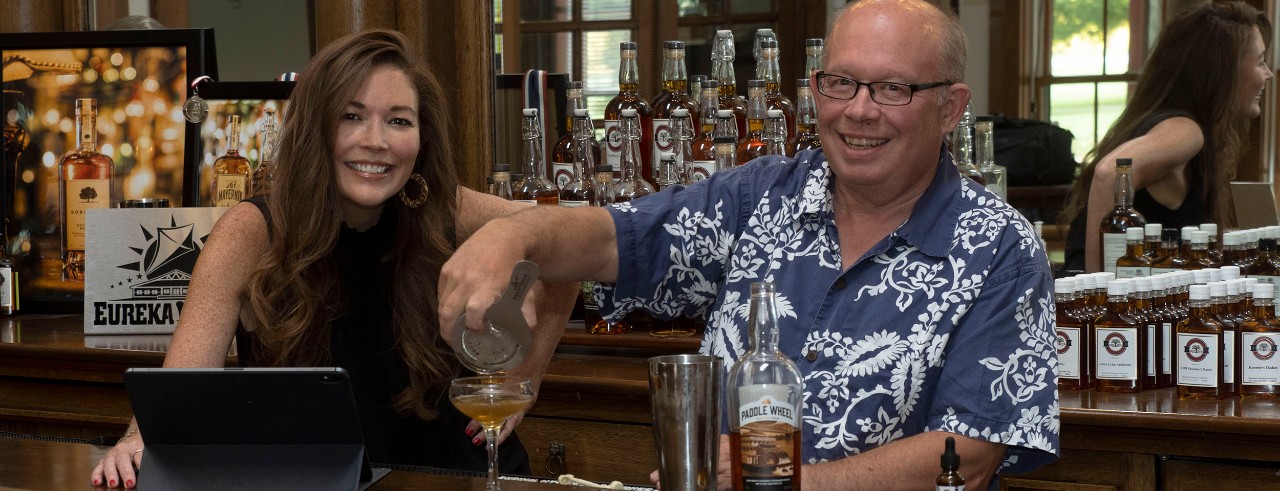 Woman and man business partners behind a bar smiling, man pouring drink, woman with tablet.