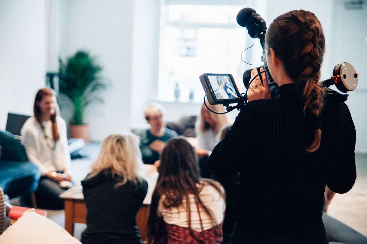 student using a video camera to conduct an interview of a group of women in a circle at a casual living room setting