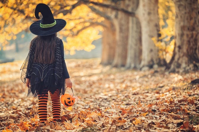 a photo of a child in a halloween costume by some trees with autumn leaves on the ground