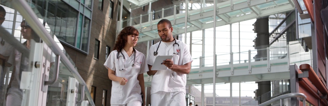 a young white woman and young black man in scrubs walk and talk in a glass-filled atrium