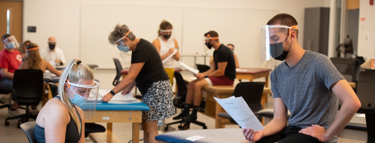 Students sitting at a table wearing masks talking to a masked instructor