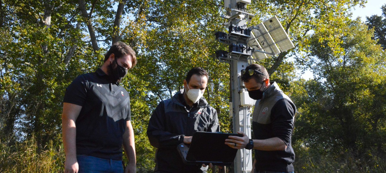 Three people wearing face masks study a laptop in front of a steel pylon mounted with electronic equipment surrounded by trees.
