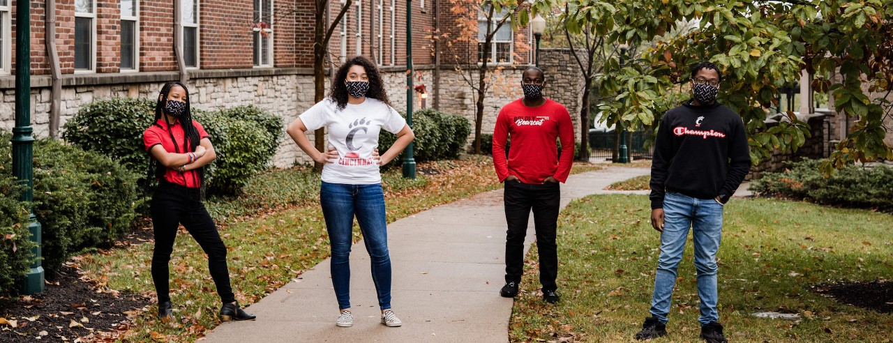 4 students in UC gear pose outside on campus
