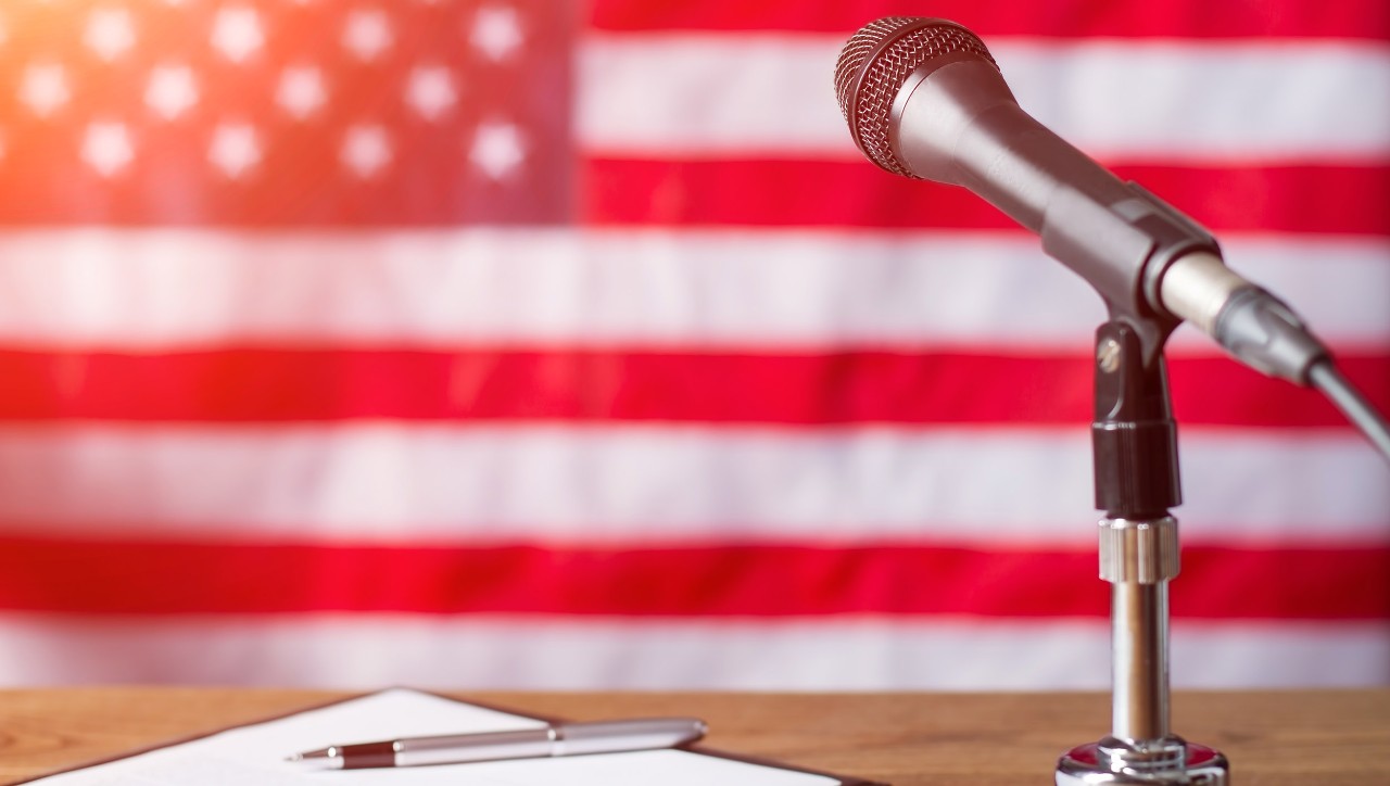 A microphone and podium in the foreground, with an American flag in the background.