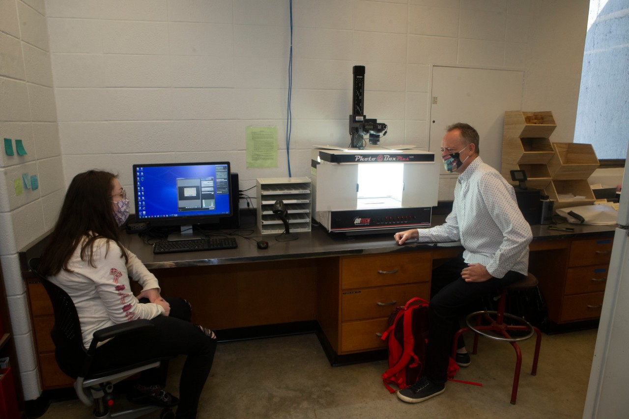 Two people in face masks sit at a lab bench in front of a photo lightbox.