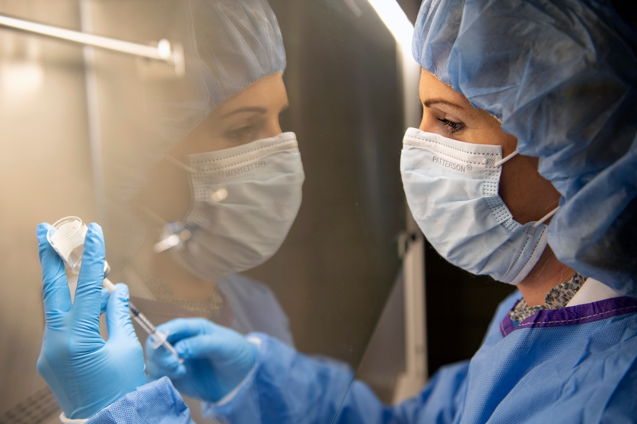 A researcher wearing a mask that is reflected in a laboratory window as she holds a vaccine vial