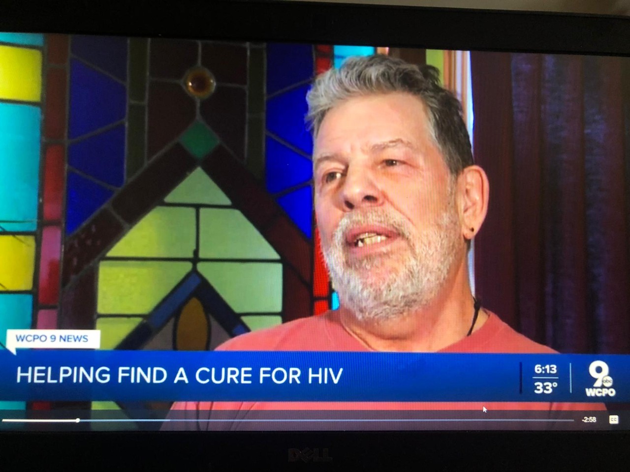 A screen shot from a TV broadcast with a bearded man talking and the headline says helping find a cure for HIV
