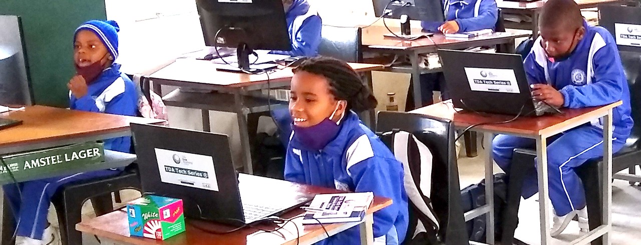 Students in blue uniformssit at desks in a classroom, looking at laptops and monitors; they wear masks pulled down to their chins; in the foreground, a girl smiles widely; in the middle ground are two boys .