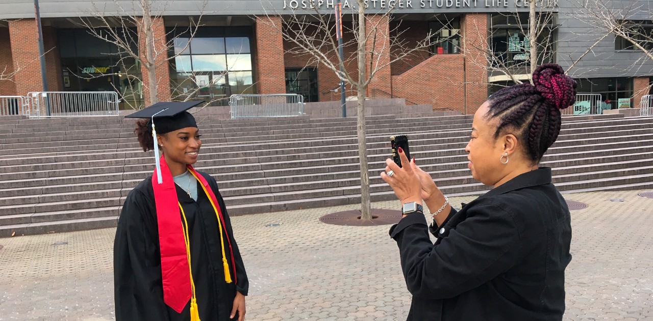 A UC student in her cap and gown poses for a photo that her mother takes on UC's Uptown Campus.