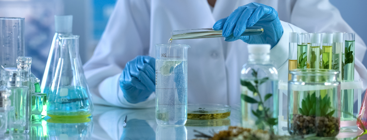 a person in a white lab coat and blue gloves stands at a lab bench pouring liquid into a beaker. There are several glass containers on the bench containing plants and liquids