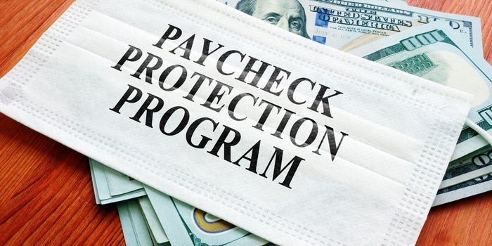 Picture of a face mask with the words "Payroll Protection Program" written across the front. The mask is sitting atop a pile of $100 bills. 