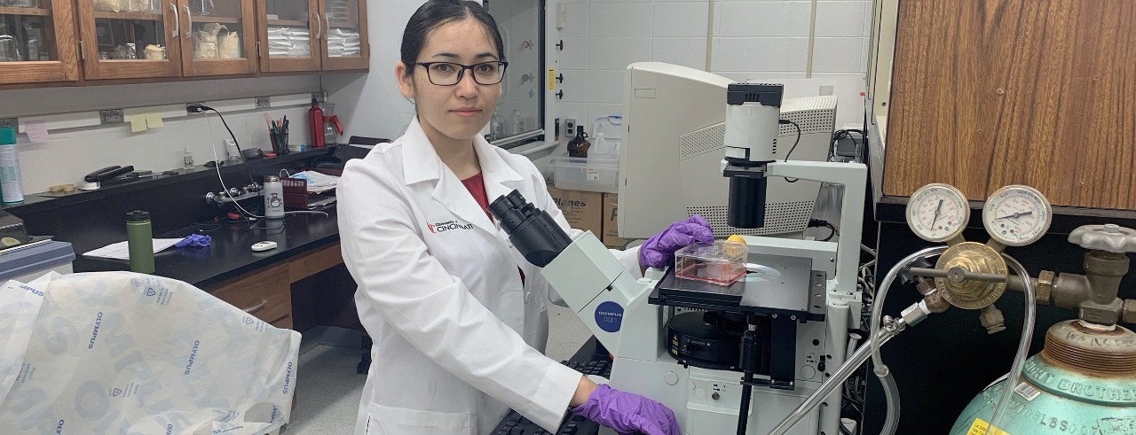 A young woman wearing a lab coat and gloves stands at a microscope in a lab.