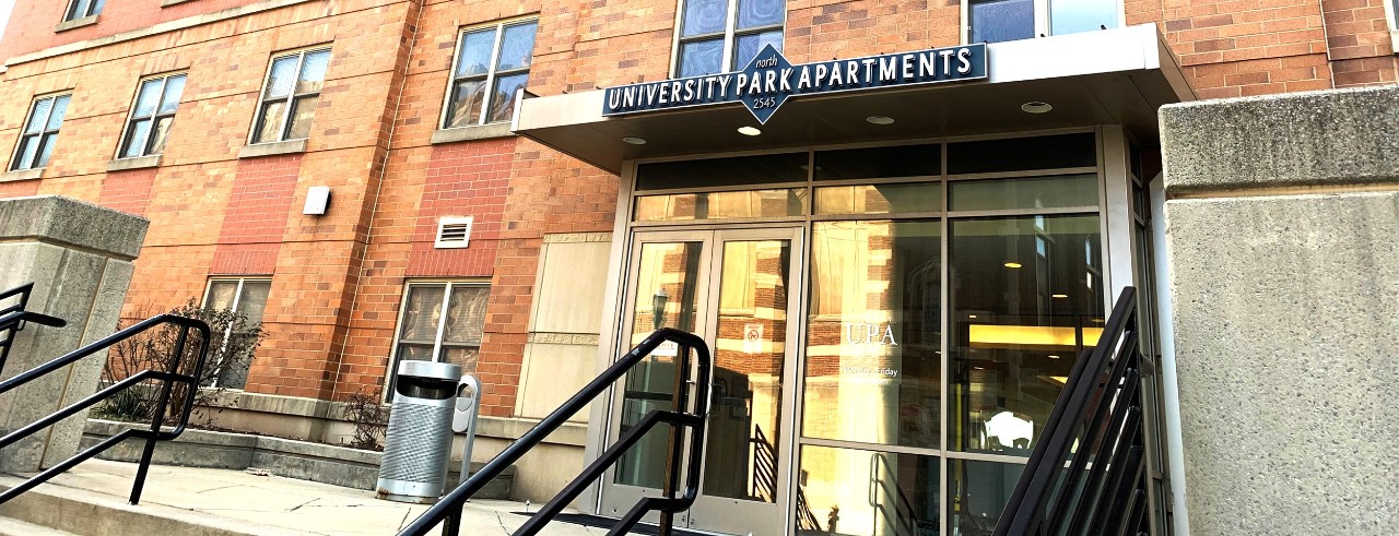 Doorway to an orange brick building with concrete paver stairs, black railings and glass doors topped with an overhang with a sign that says "University Park Apartments"