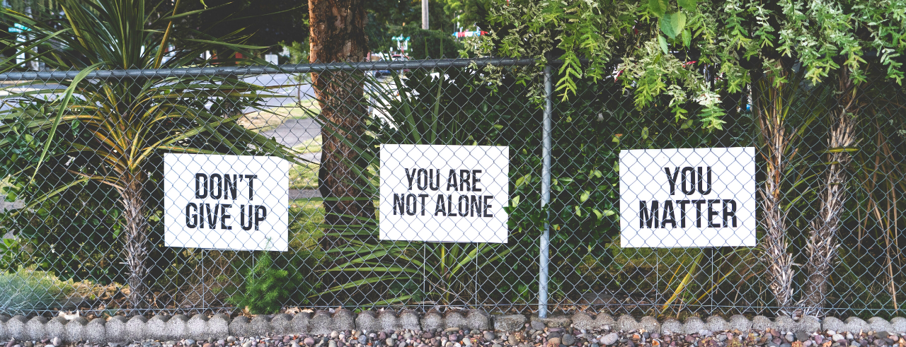 a chainlink fenceline with signs that read, "don't give up; you are not alone; you matter", s trees and bushes behind
