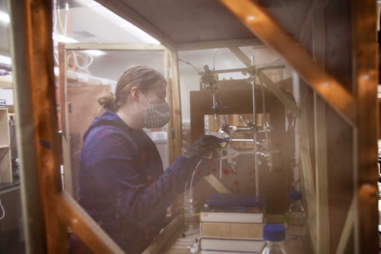 A student is seen through a mesh screen at a chemistry work station in a lab.