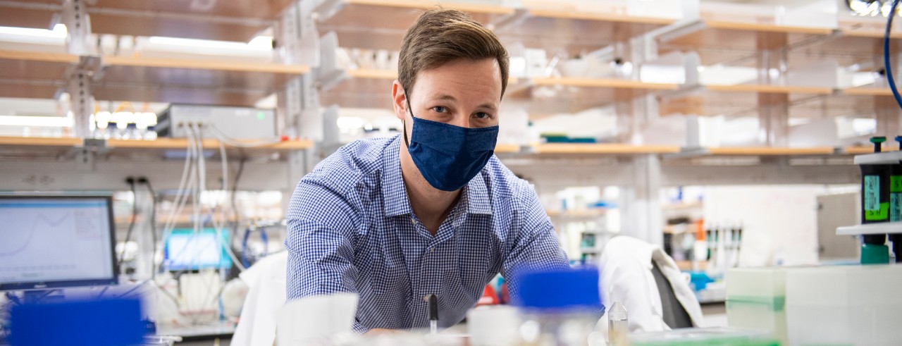UC student Andrew Eisenhart, wearing a face masks, peers a shelf of chemistry equipment in a lab.
