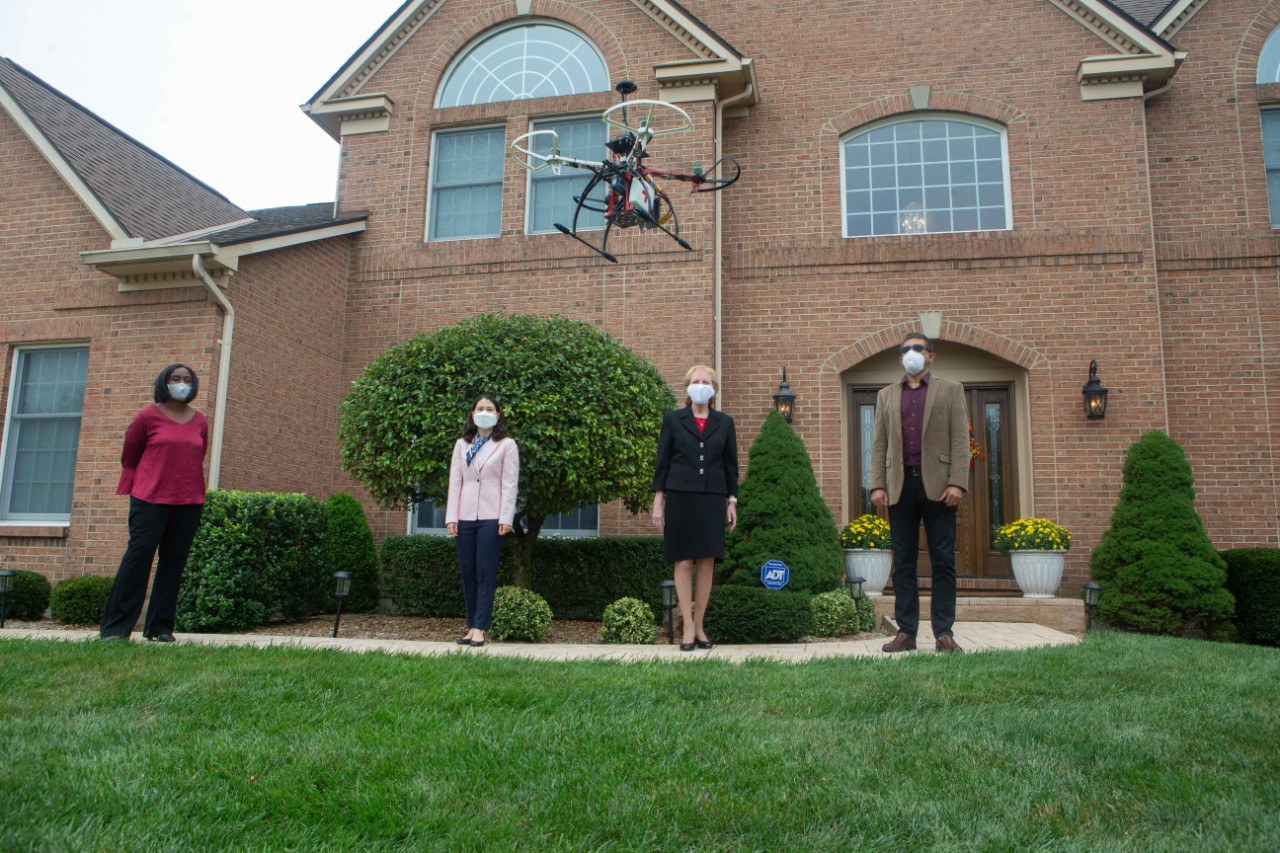 Four people socially distanced and in face masks stand outside a home with a drone hovering over them. 