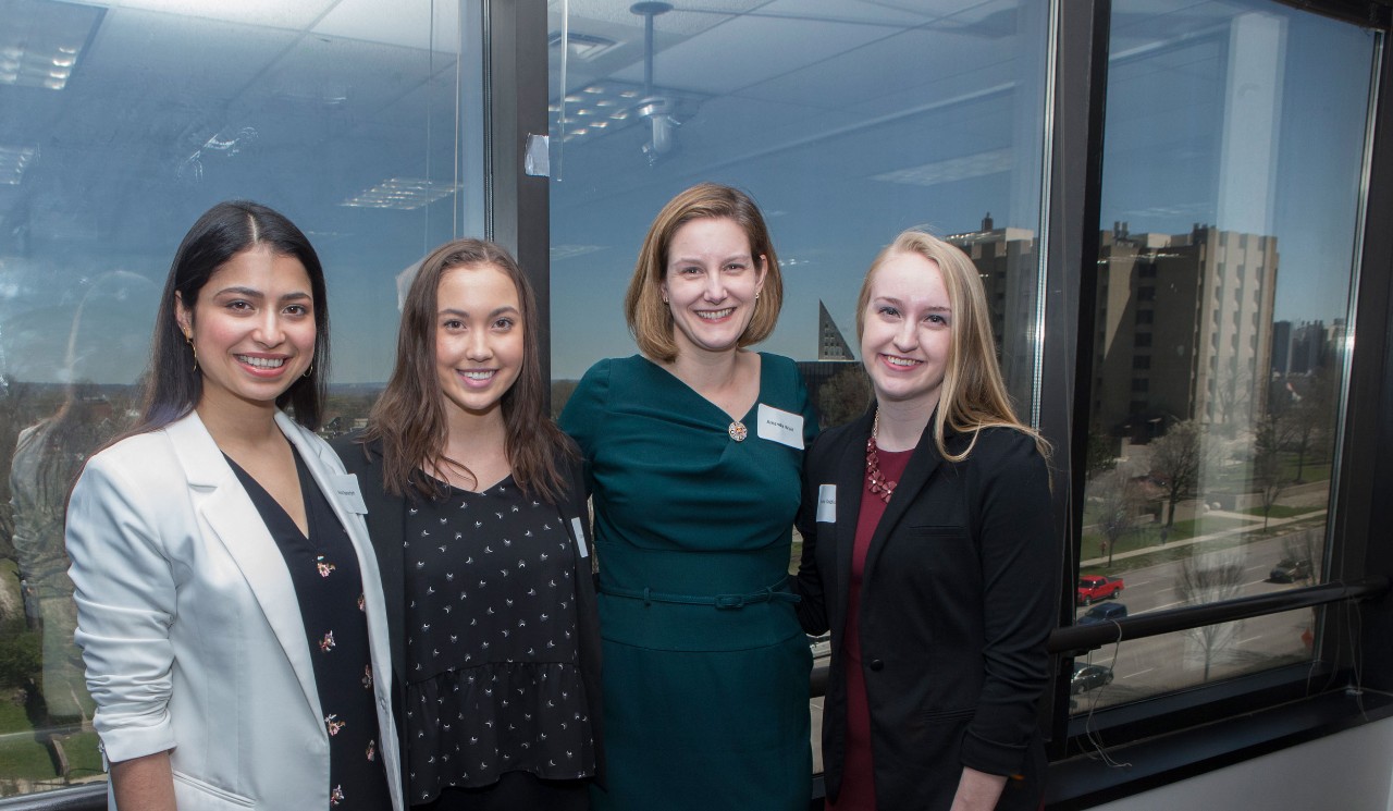 four women in professional clothing stand close to one another and smile for a group photo. from left to right: woman with dark hair and white blazer, woman with dark hair and black dress, Amanda wait with chin-length hair and dark green dress, and woman with blonde hair and black blazer