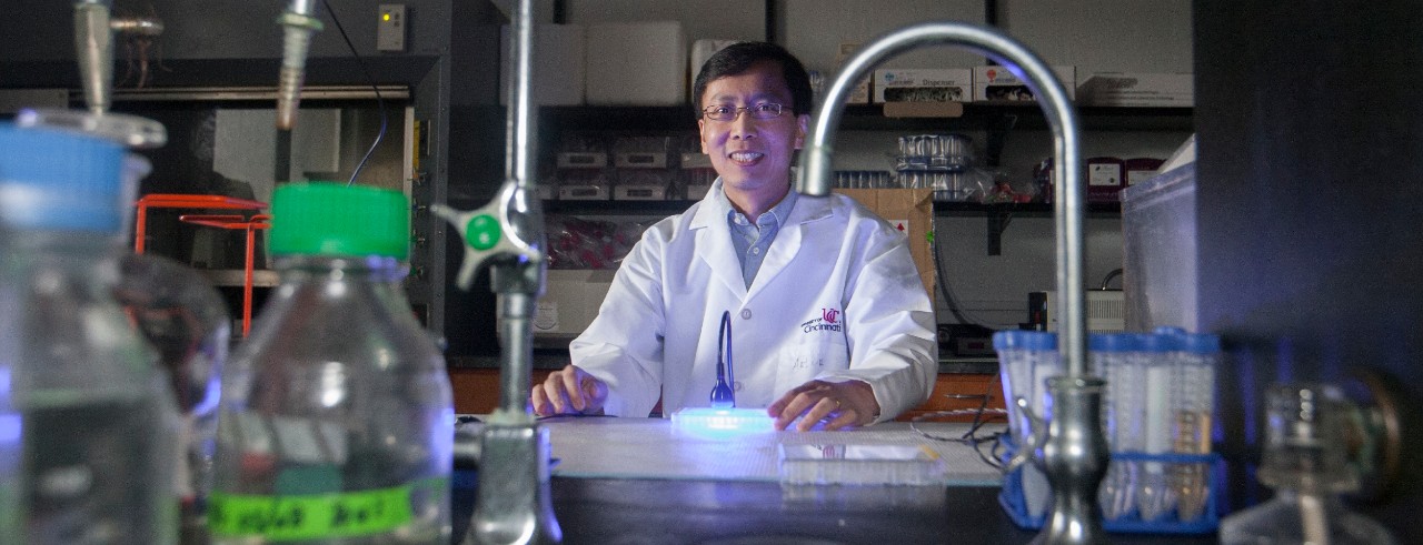 A man in a lab coat stands at a chemistry lab bench with a blue light shining down on a petri dish.