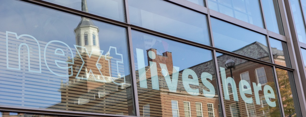 McMicken hall reflected in windows with "Next Lives Here" sign on them
