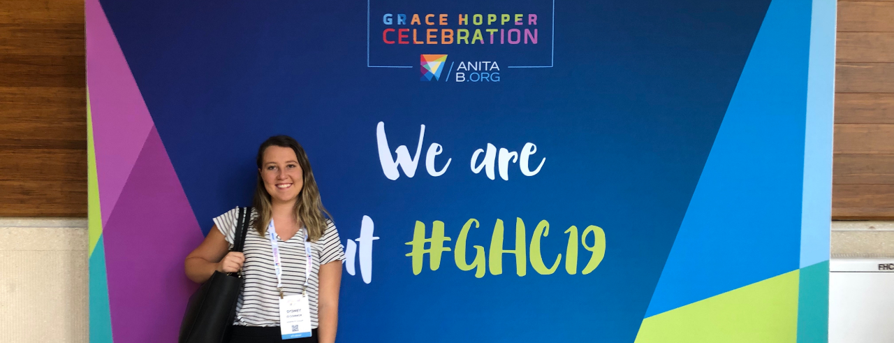 a young woman in business casual wear stands in front of a banner for the Grace Hopper celebration 2019 conference