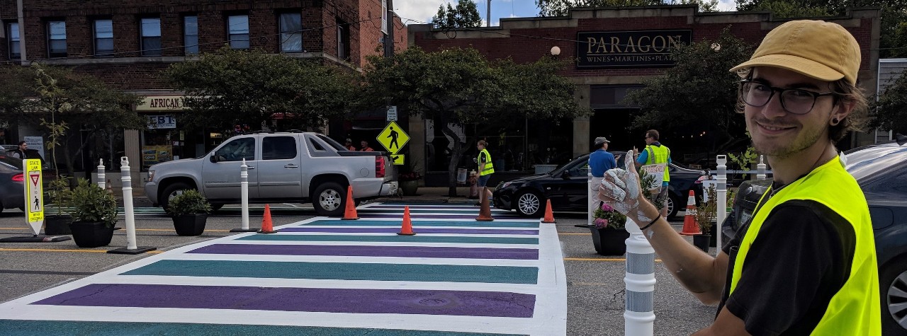 young man in safety vest gestures to a wide crosswalk painted white, purple and green