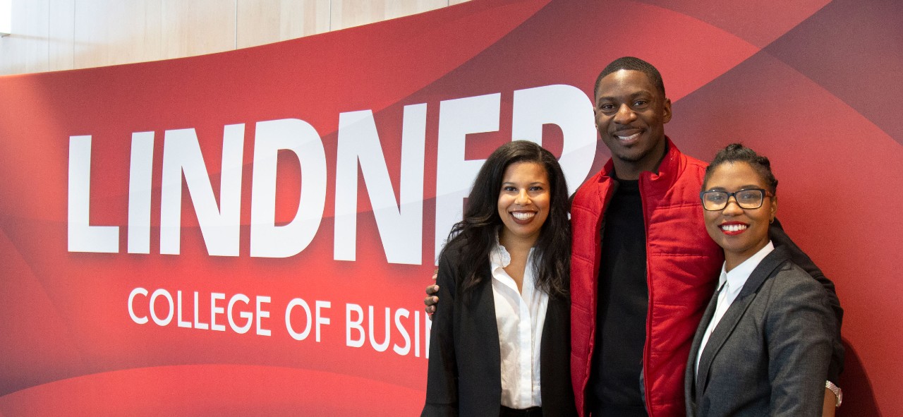 A Back man wearing a red vest stands between two Black women wearing dark suits and white blouses, all smiling and huddled close in front of a red backdrop that says LINDNER