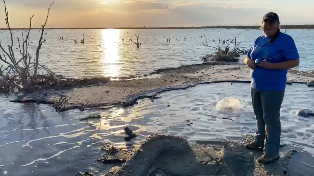 UC associate professor Amy Townsend-Small stands in front of a small lake created by a bubbling oil well and the setting sun behind her.