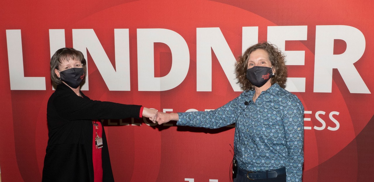 A woman in a black sweater fist bumps with another woman in a blue patterned blouse in front of a red banner with LINDNER in white