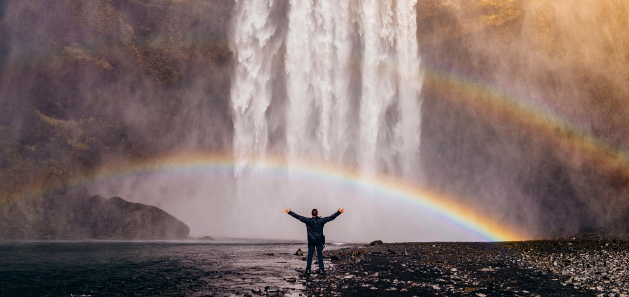 A person stands with his arms outstretched under a waterfall with two rainbows in the mist.
