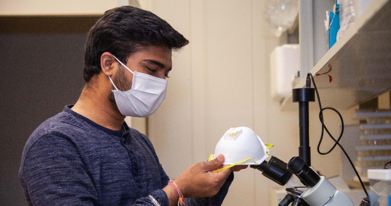 A UC student wearing a face mask holds another mask in a UC lab.
