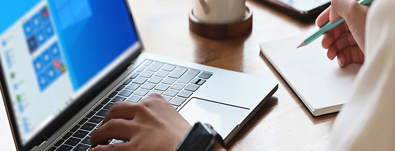 A closeup of a hand on a keyboard and another holding a pencil over a notebook.