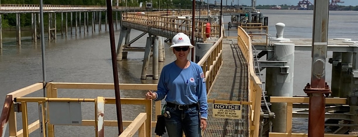 Female college student wearing work clothes and a hard hat stands on a dock near water