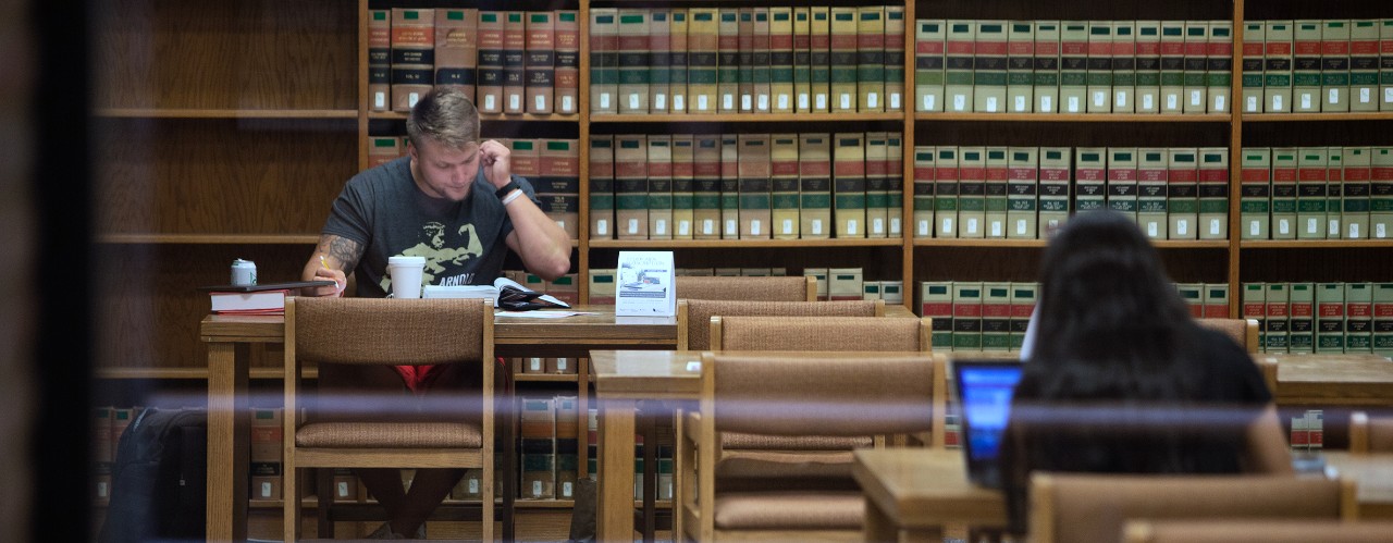 Man looks down at book and writes on paper while sitting at a library table.