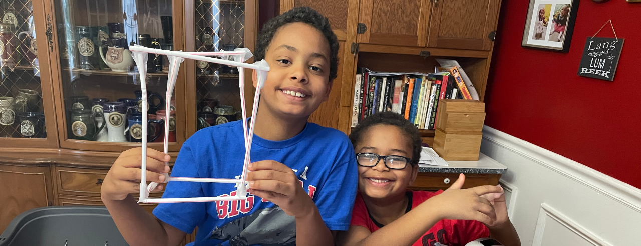 two children sit smiling at a table, one is holding a structure made out of drinking straws and tape