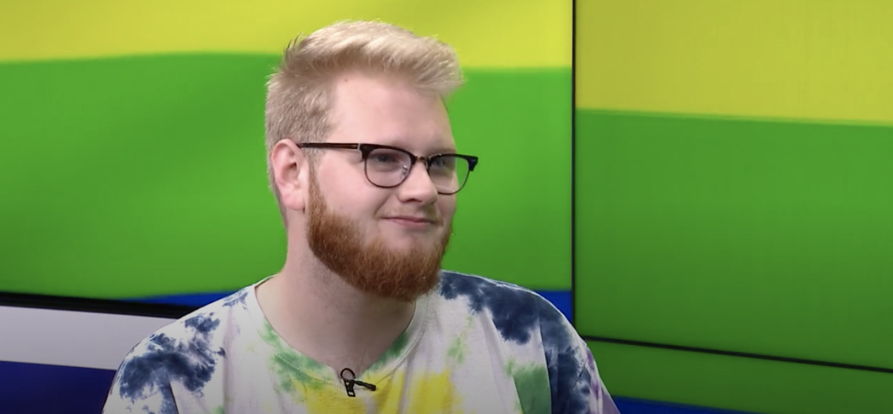 A man in a tie dye shirt sits with a green and yellow background behind him.