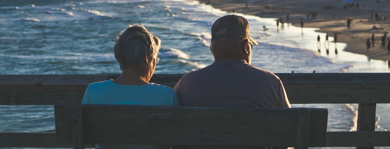 An elderly couple sits on a bench looking out at a body of water