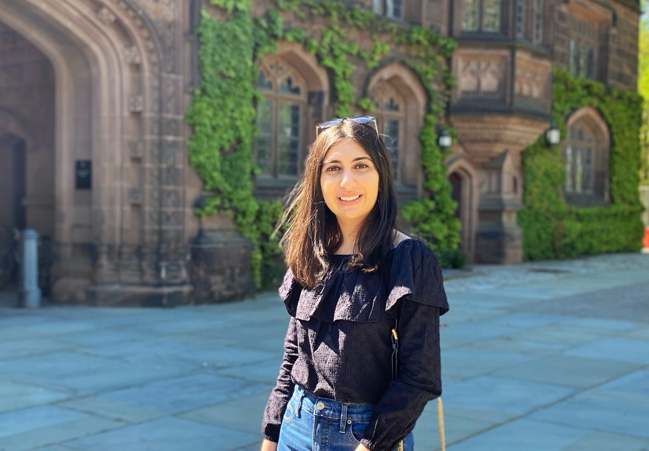 A woman in a black shirt and jeans stands with a building in the background.