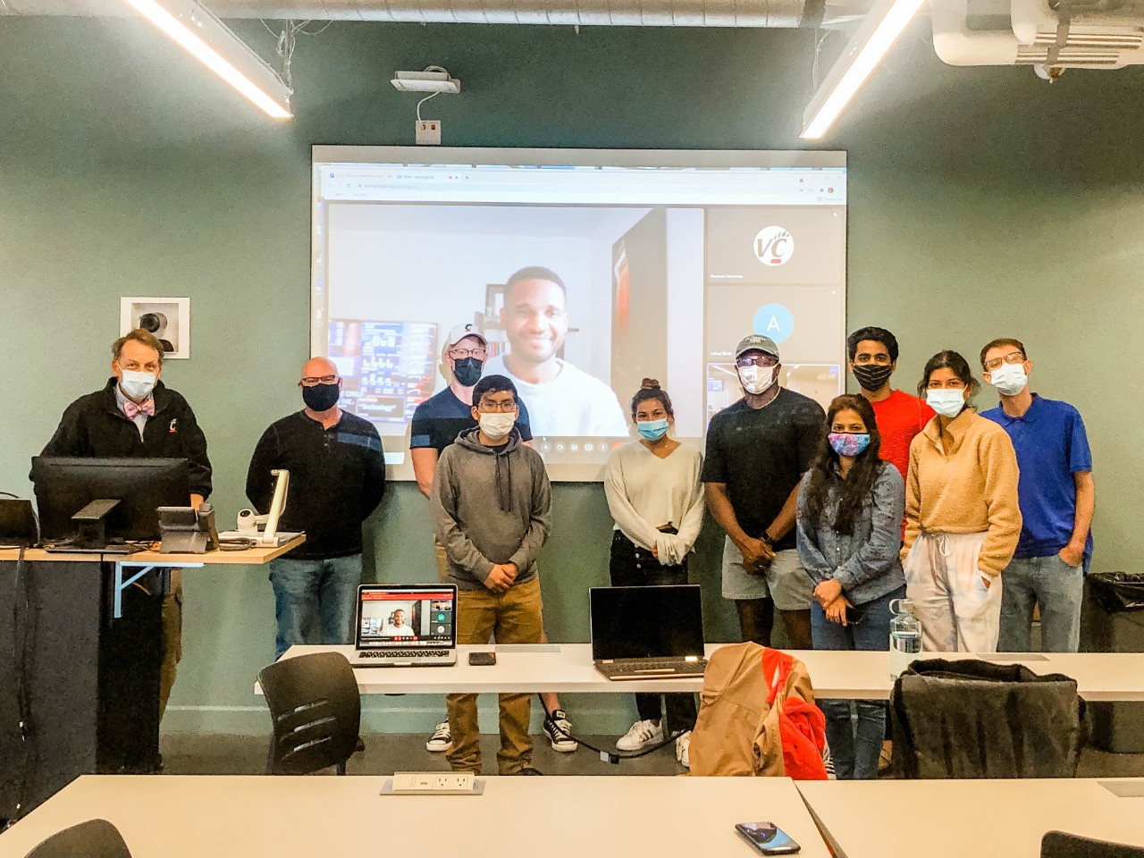 A speaker on a screen smiles as a group of organization members pose in front of him.