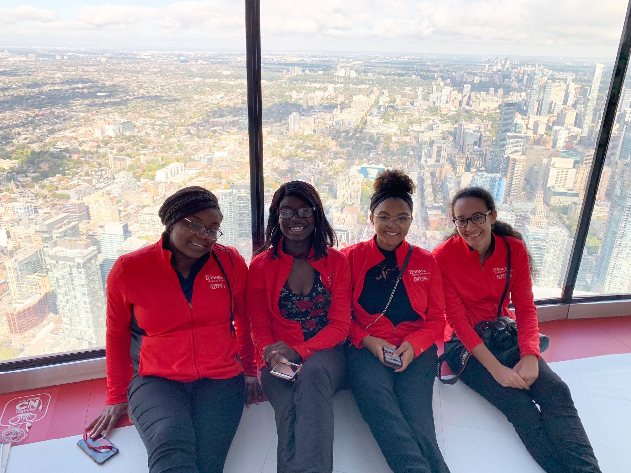 Four students in matching red jackets smile for the camera with a city skyline in the background.