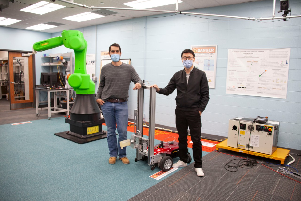 Two students in face masks stand next to a wheeled robot in a robotics lab.