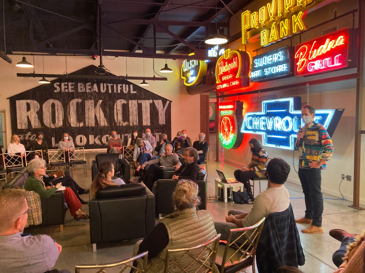 Nathan Morehouse makes introductory remarks to guest speakers gathered in a circle in front of the neon signs of the American Sign Museum.