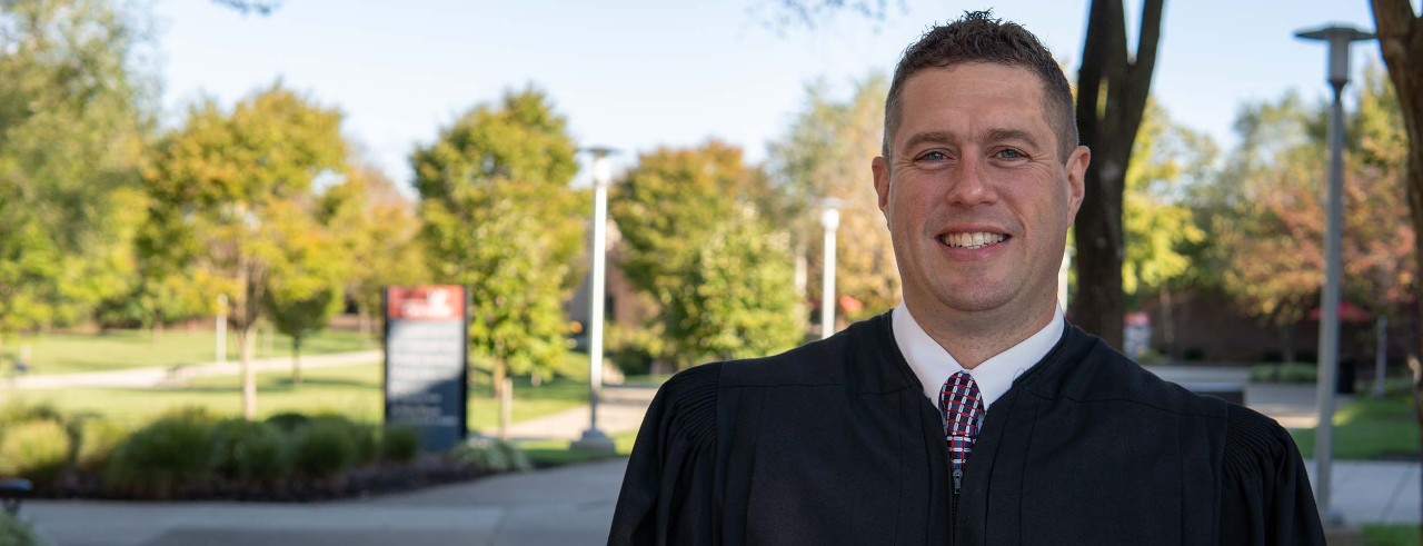 Nathan Little in judge robes smiling on UC Clermont campus.