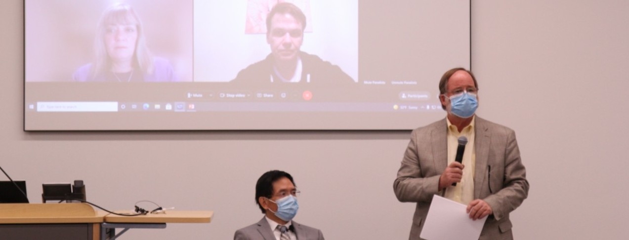 A man in a beige jacket speaks before a crowd during a forum.