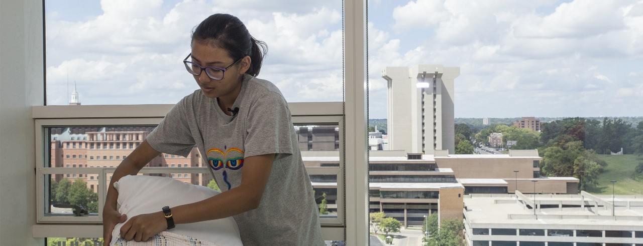A student puts a case on a pillow in her dorm room, with a view of the UC campus through the window behind her.