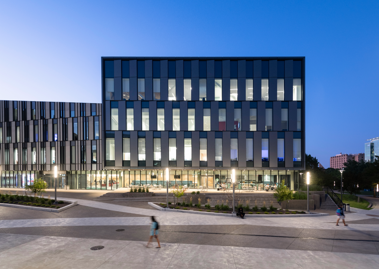 At night, a building is lit up by its many windows as people walk by.