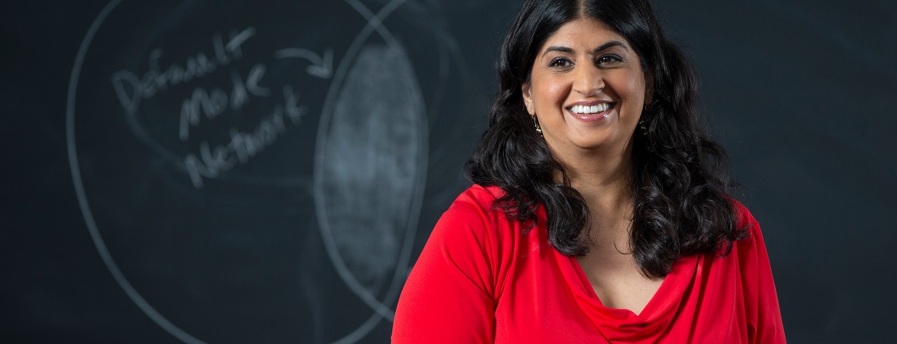 Meera Rastogi wearing a red dress and standing in front of a black board with symbols in chalk 