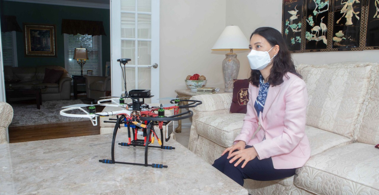 A woman in a face mask sits on a couch in front of a drone sitting on a coffee table in front of her.